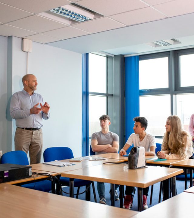 A mid adult man teaches an engineering class to a group of students. A female tutor stands and observes the lesson.